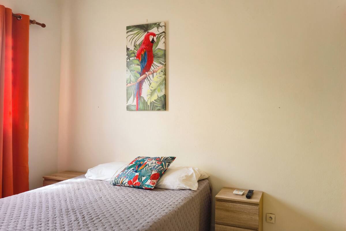 Ein Schlafzimmer mit einem Bett, einem Nachttisch, einem Papageienbild und einem Fenster mit Fensterläden.