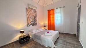 A bedroom with a bed, nightstands, a wall-mounted air conditioner, and a large window with orange curtains. - Domaine de May (Saint-François)