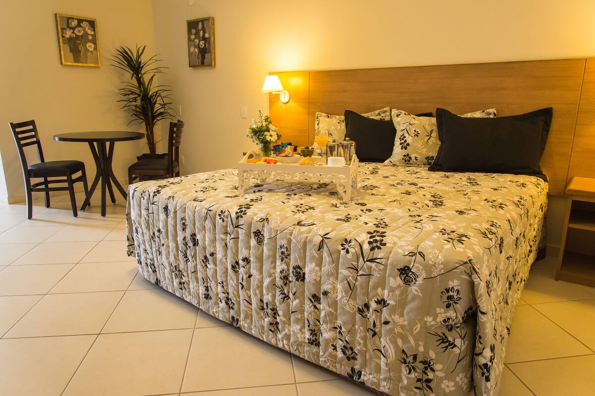 A hotel room with a large bed, a bedside table with a lamp, a small table with a plant, and a chair.