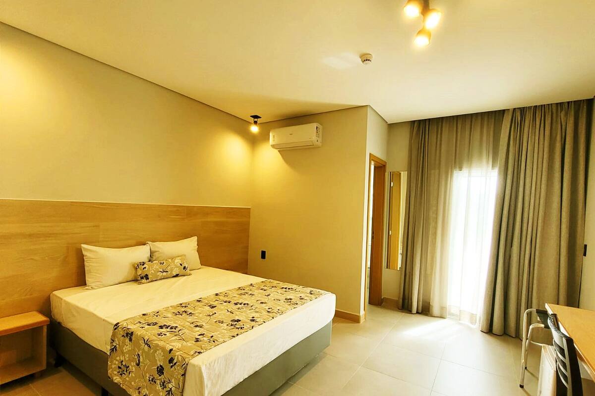 A hotel room with a large bed, a wooden headboard, a desk, and a window with curtains.