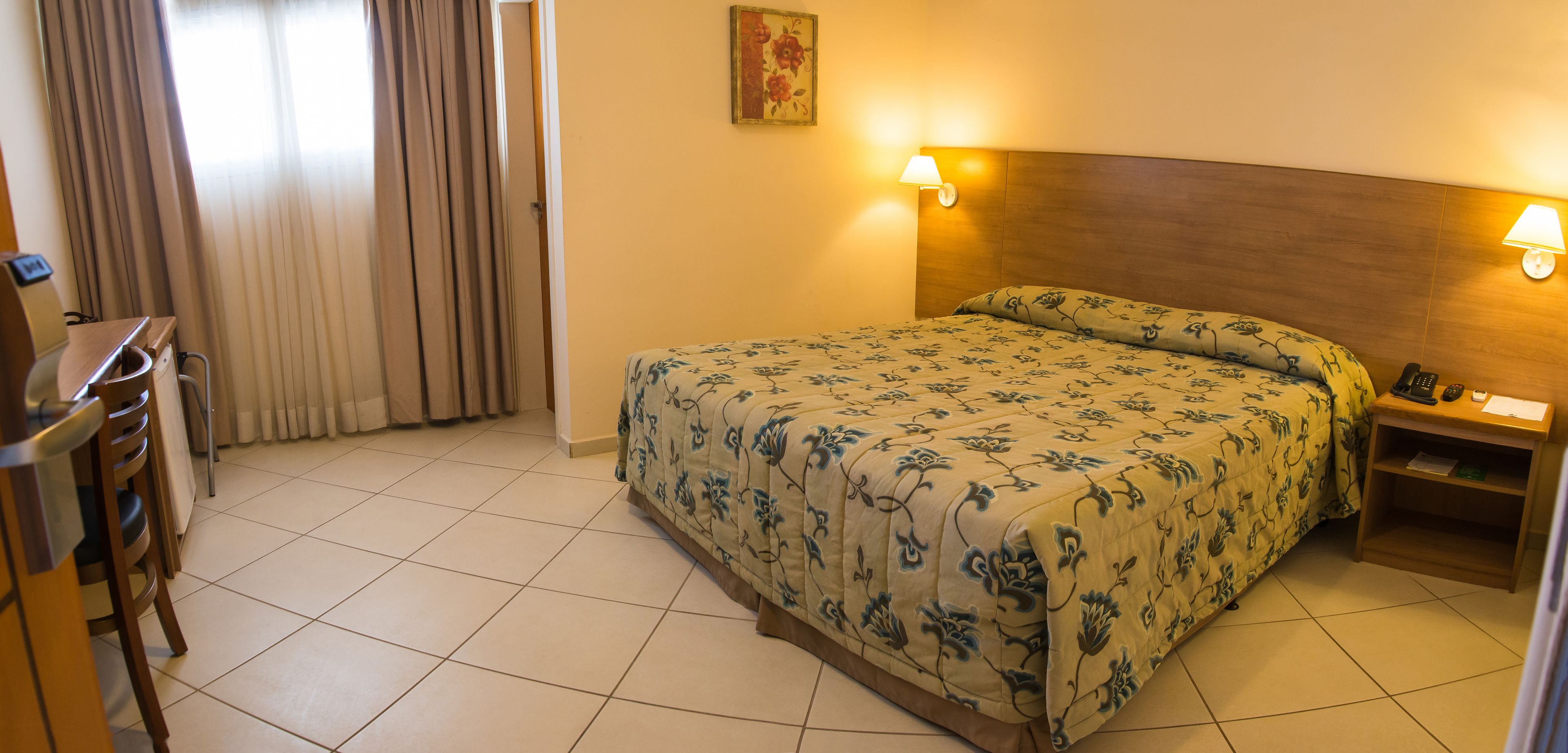 A hotel room with a bed, a desk, a chair, a nightstand, a lamp, and a telephone.