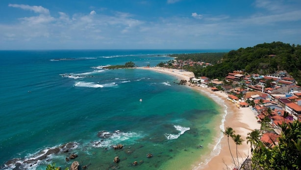 Un pueblo costero con playa, aguas cristalinas y un paisaje montañoso al fondo.
