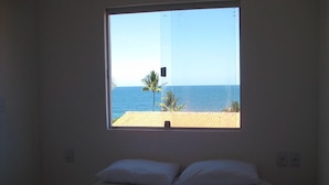 Un dormitorio con un amplio ventanal que da a una playa, una cama con ropa de cama blanca y vista al océano.