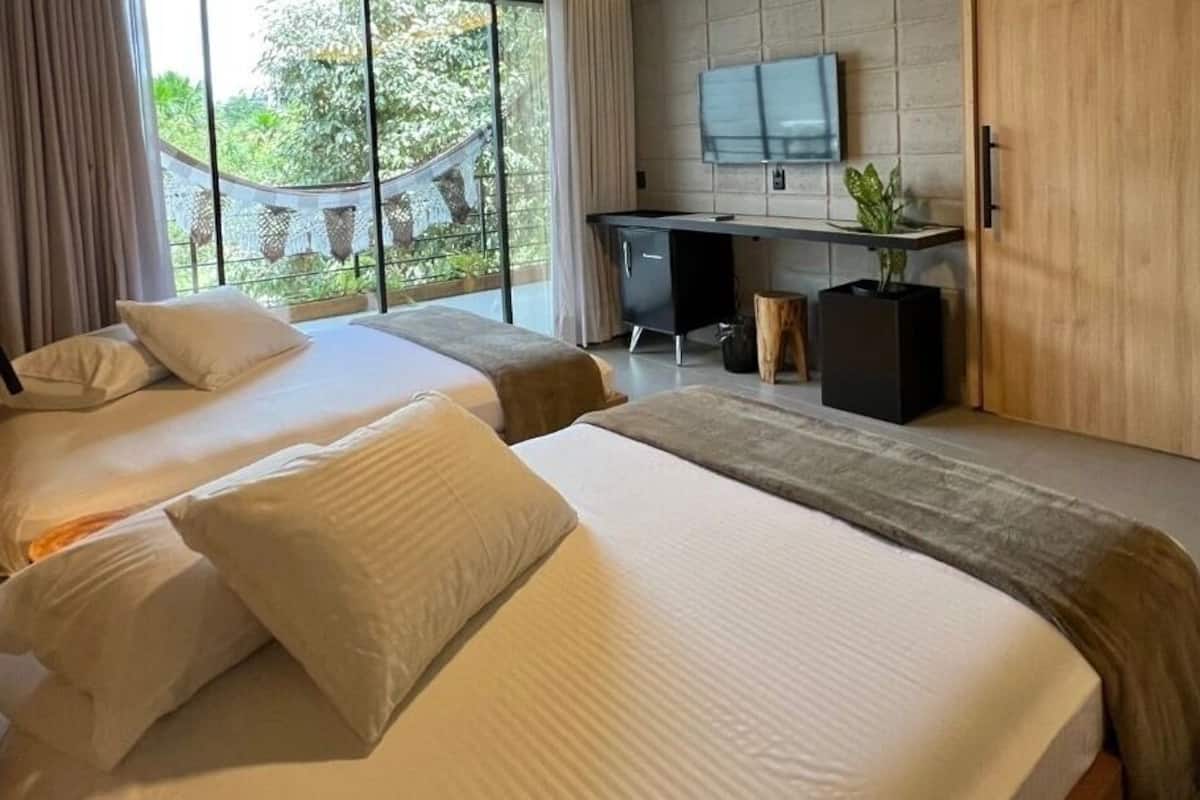 A hotel room with two beds, a desk, a TV, and a balcony with a view of greenery.