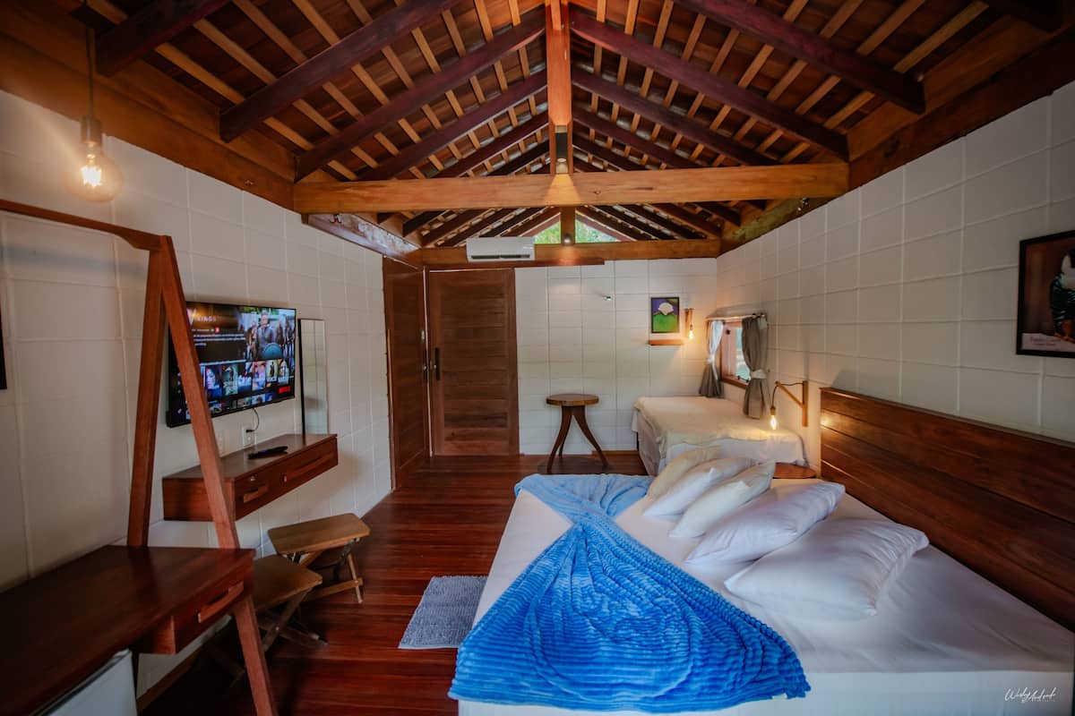 A bedroom with a wooden ceiling, a bed with white bedding, a desk with a chair, a television, and a small stool.