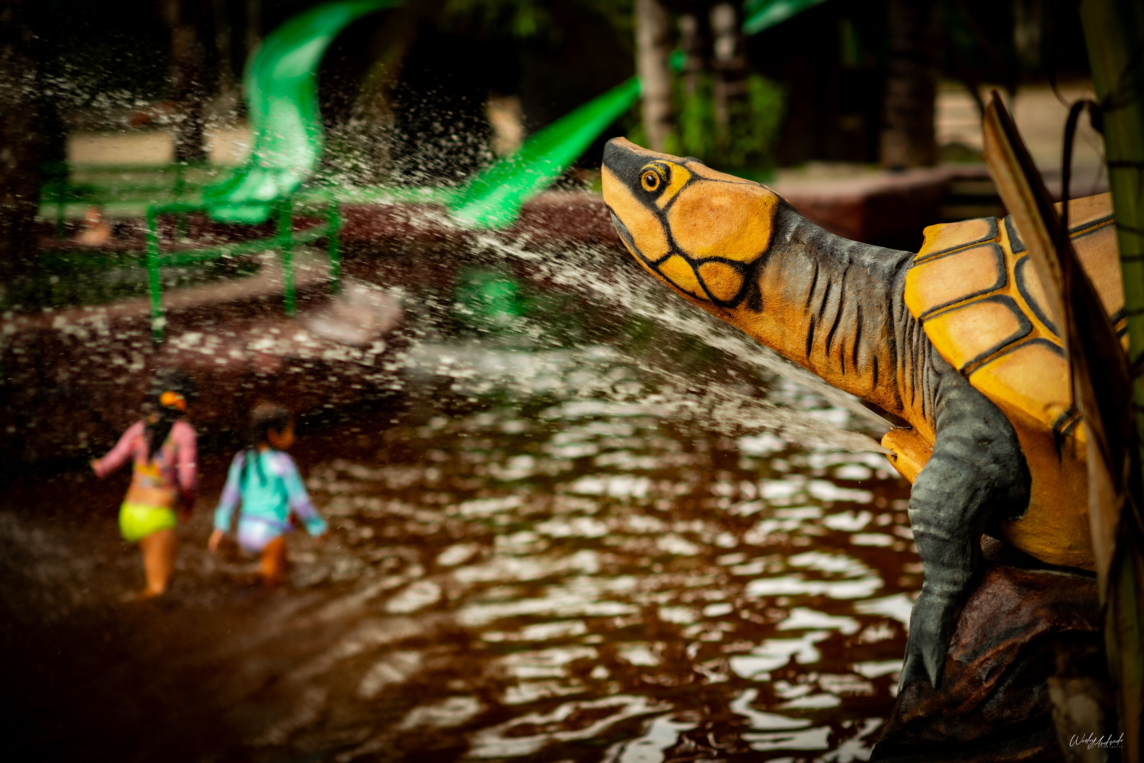 A dinosaur statue with water spraying from its mouth, a green slide, and two people in the background.