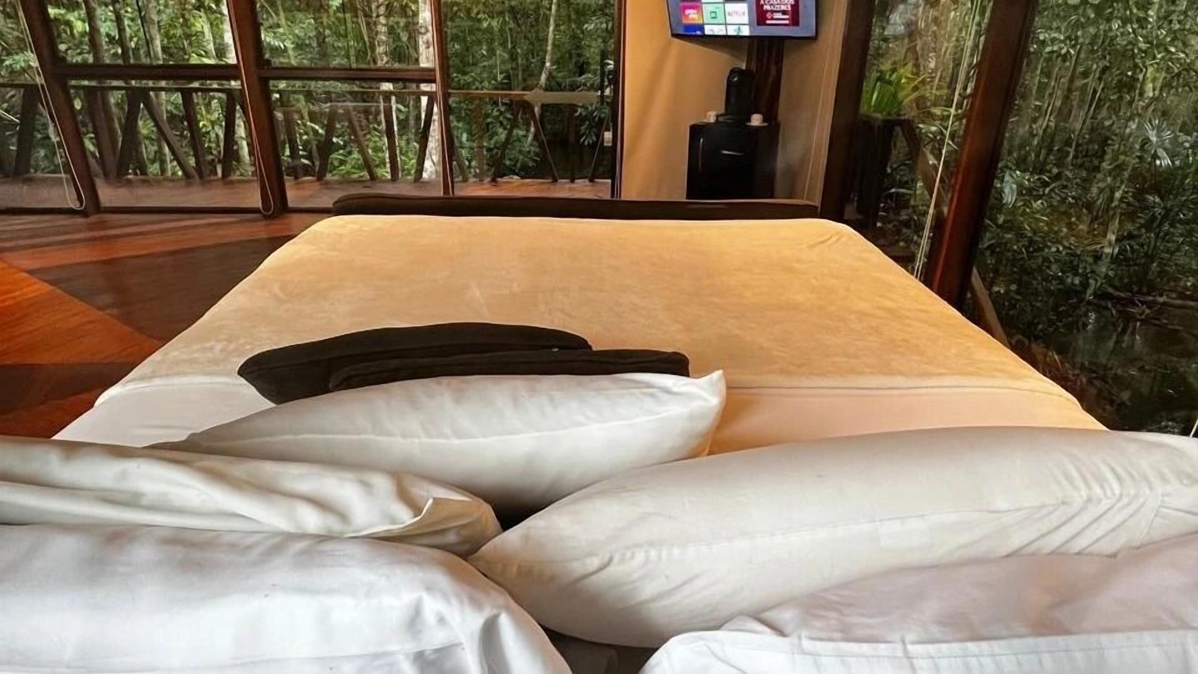 A bed with white and black pillows, a TV on the wall, and a view of a forest through large windows.