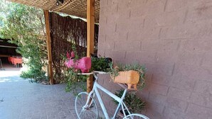 Un vélo est garé sur un patio pavé de pierres, agrémenté de plantes en pot et surmonté d’une sorte de tonnelle faite de matériaux tressés.