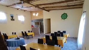 Un espace repas avec plusieurs tables et chaises, une cuisine à l’arrière-plan et un plafond rustique.