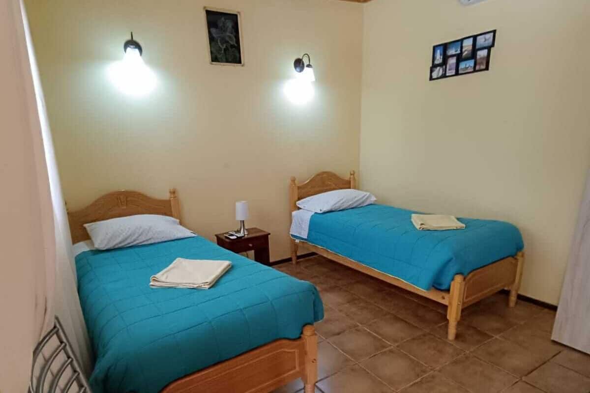 Une chambre avec deux lits en bois, chacun recouvert d’une couverture bleue et agrémenté d’oreillers blancs. Une petite table de chevet avec une lampe se trouve à côté de chaque lit. La chambre a un sol carrelé et un plafond en bois avec des poutres apparentes.