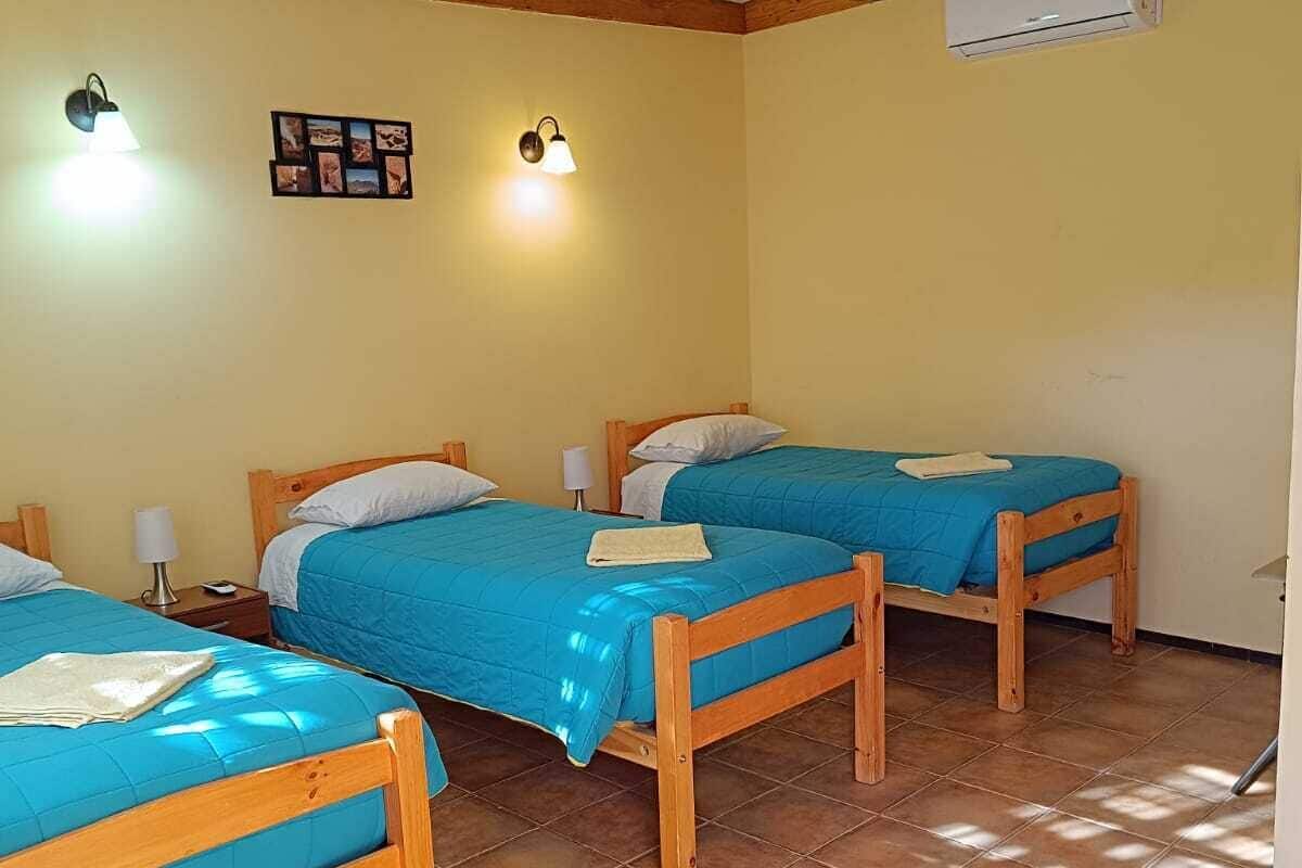 Une chambre avec trois lits en bois, chacun recouvert d’une couverture bleue et agrémenté d’oreillers blancs. On y trouve des lampes fixées au mur et un appareil de climatisation.