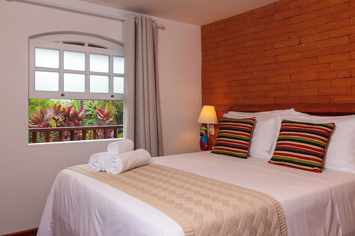 A neatly made bed with a white comforter and a beige throw blanket, a window with curtains, and a brick wall.