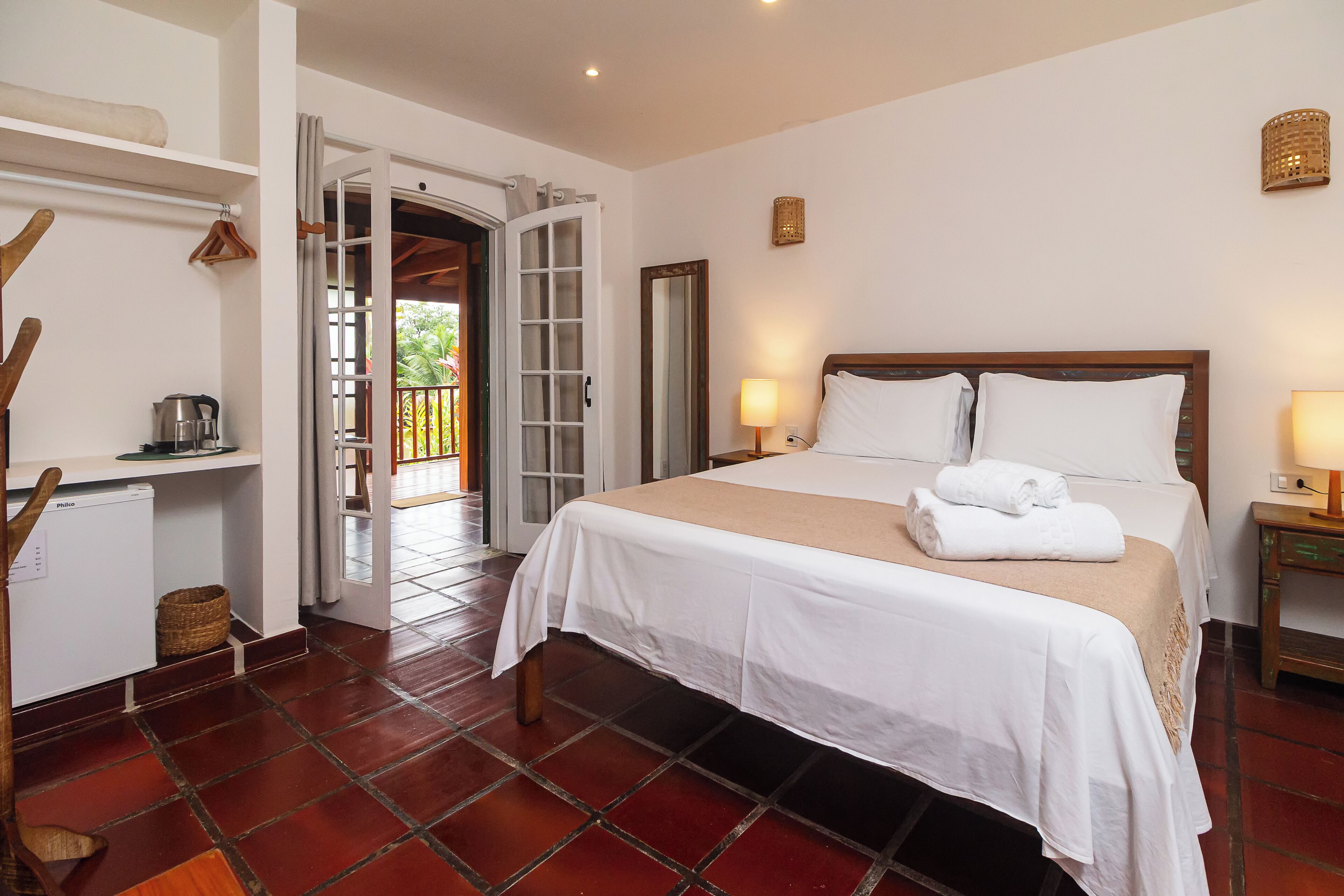 Habitación de hotel con cama, una pequeña mesa de trabajo con tetera, una cesta y una puerta que da a un espacio exterior.