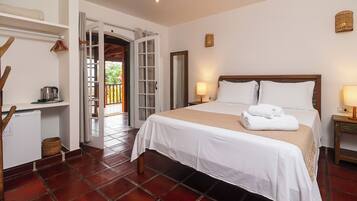 Habitación de hotel con cama, una pequeña mesa de trabajo con tetera, una cesta y una puerta que da a un espacio exterior.