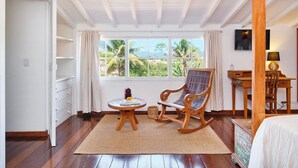 A room with a wooden floor, a rocking chair, a small round table, a bed, a TV, and a window with a view of palm trees and mountains.