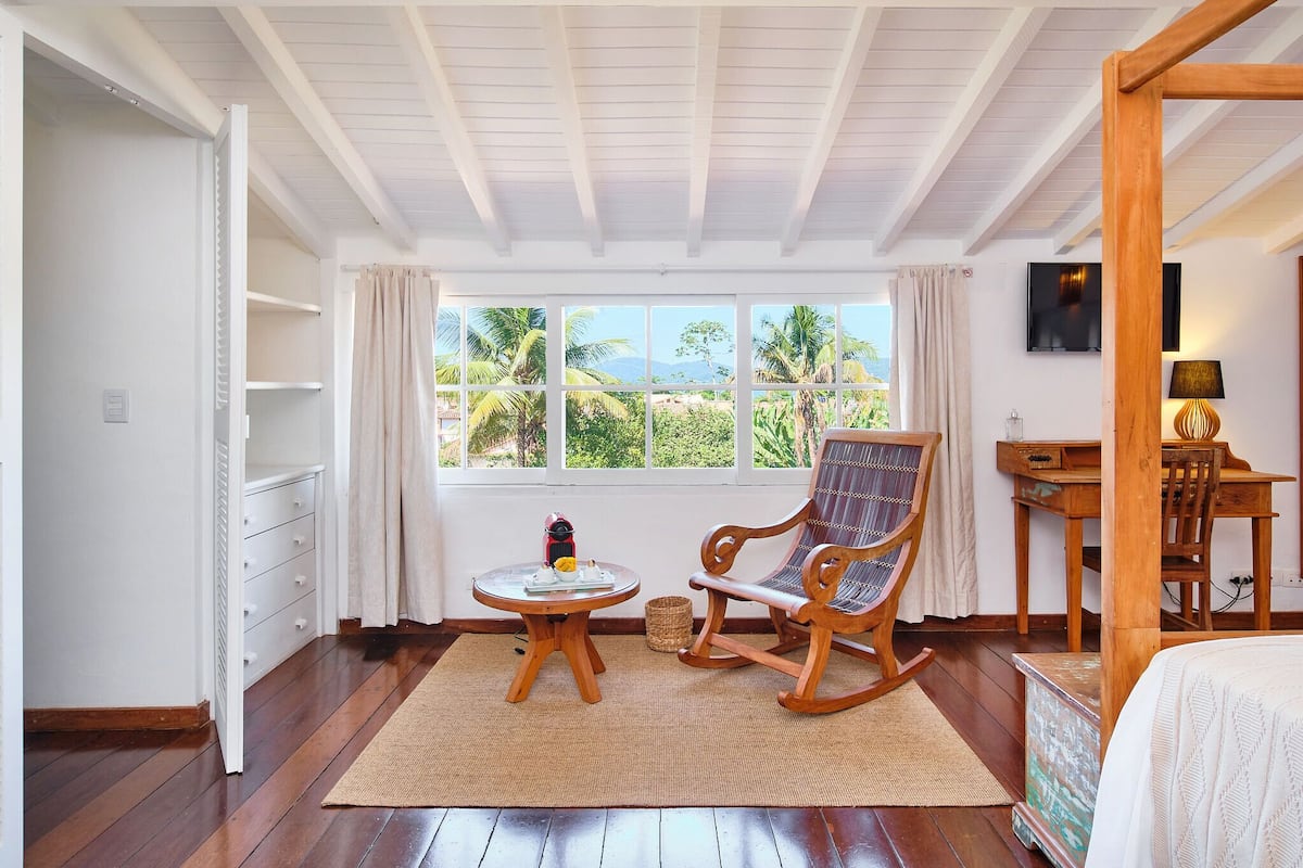 A room with a wooden floor, a rocking chair, a small round table, a bed, a TV, and a window with a view of palm trees and mountains.