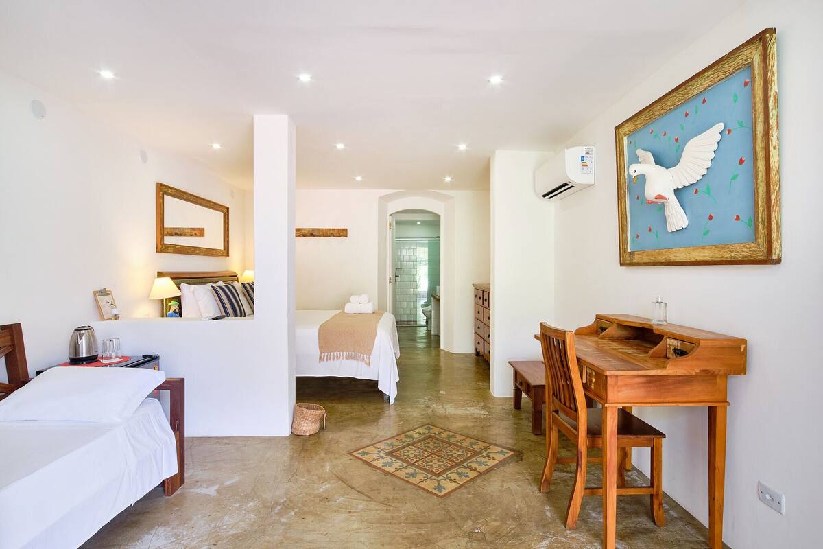 A hotel room with a bed, a wooden desk, a chair, a small table, a wall-mounted air conditioner, and a framed artwork of a dove.