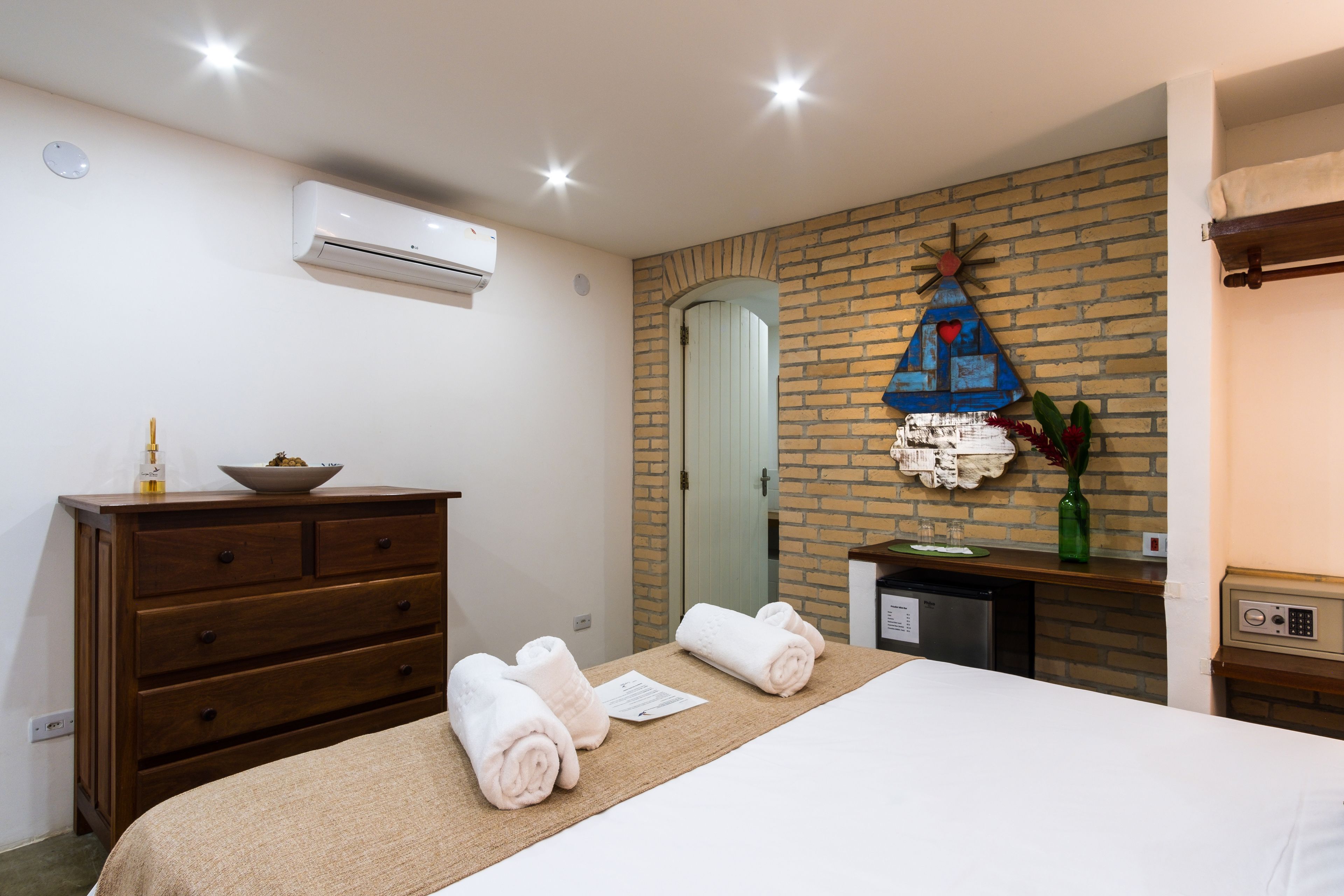 A hotel room with a bed, a wooden dresser, a small table with a vase, and a brick wall.