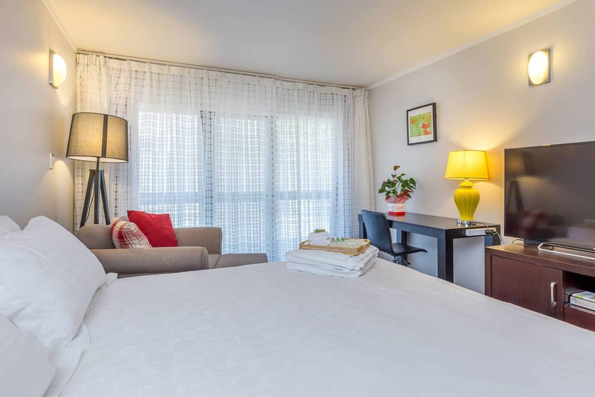 A hotel room with a large bed, a desk with a chair, a TV, a lamp, and a window with curtains.