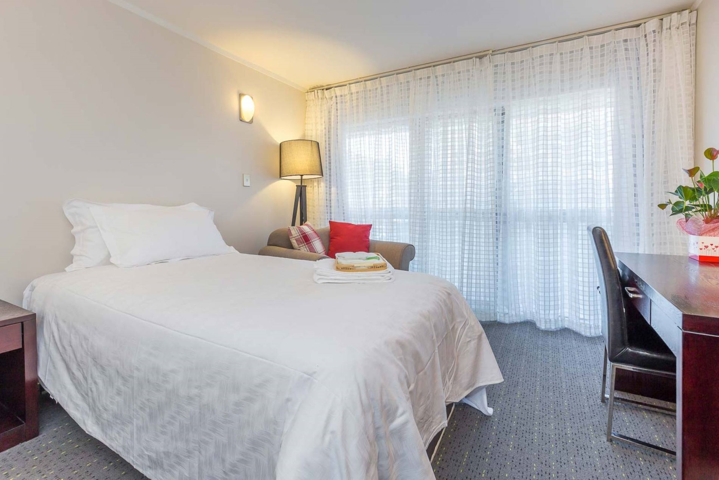 Habitación de hotel con cama, escritorio, silla, lámpara y ventana con cortinas.