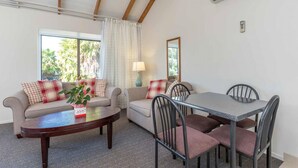 A living room with a sofa, a coffee table, and a dining table with chairs. There is a window with curtains, a lamp, and an air conditioning unit on the wall. - Anzac Court Motel (Auckland)