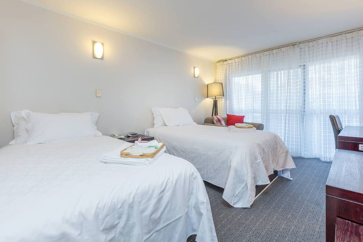 A hotel room with two beds, a desk, and a chair. There are two bedside tables with lamps and a window with curtains.
