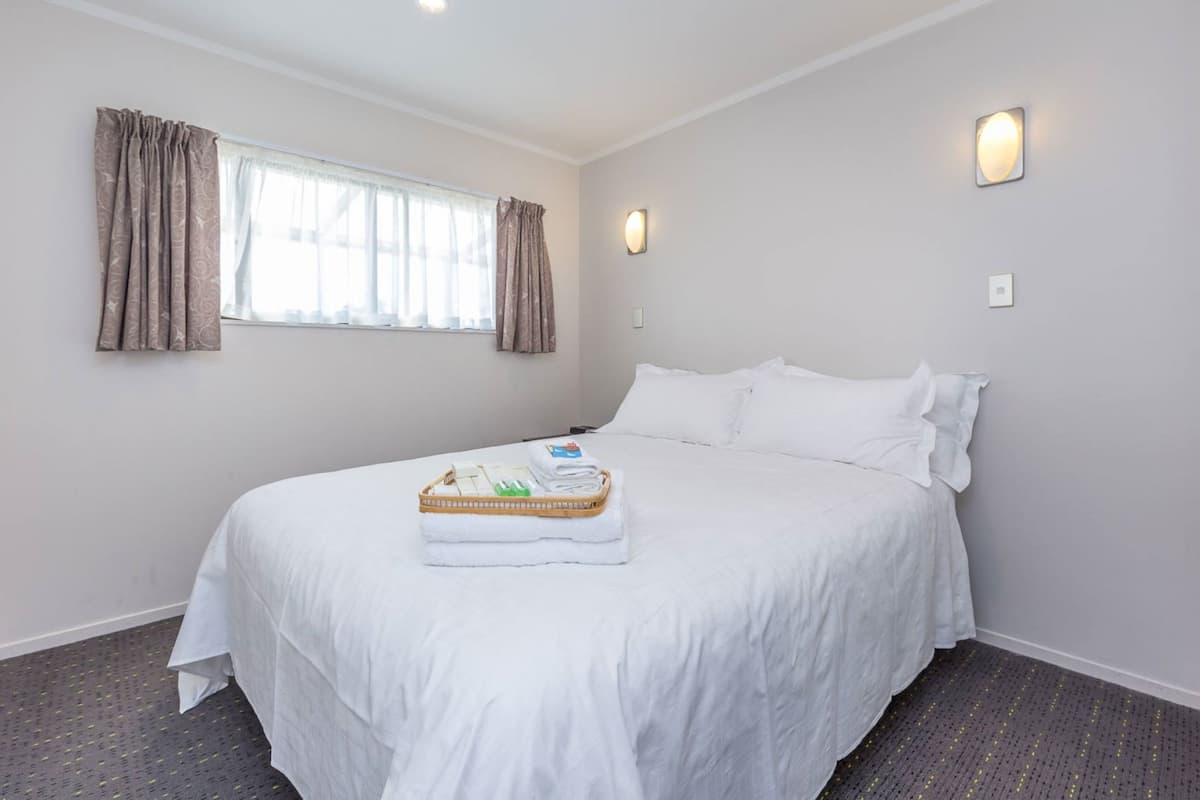 A neatly made bed with a white comforter and pillows, a window with curtains, and a wall-mounted light fixture.