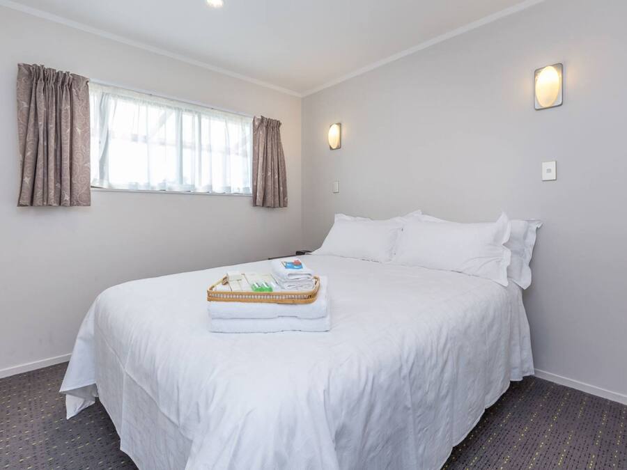 A neatly made bed with a white comforter and pillows, a window with curtains, and a wall-mounted light fixture.
