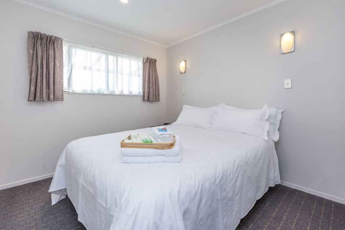 A hotel room with a large bed, two wall-mounted light fixtures, a window with curtains, and a small table with a tray of toiletries.