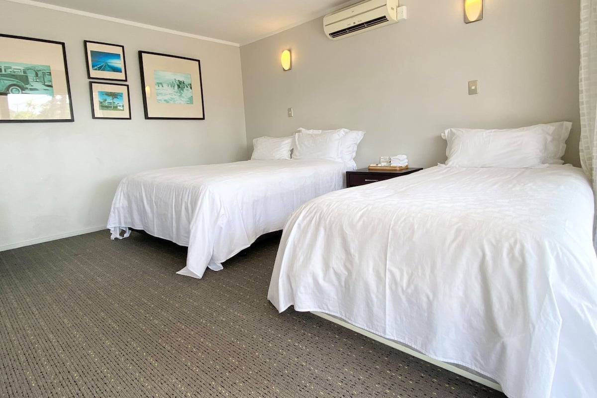 A hotel room with two single beds, a small bedside table, a wall-mounted air conditioner, and three framed pictures on the wall.