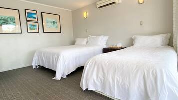 A hotel room with two single beds, a small bedside table, a wall-mounted air conditioner, and three framed pictures on the wall.