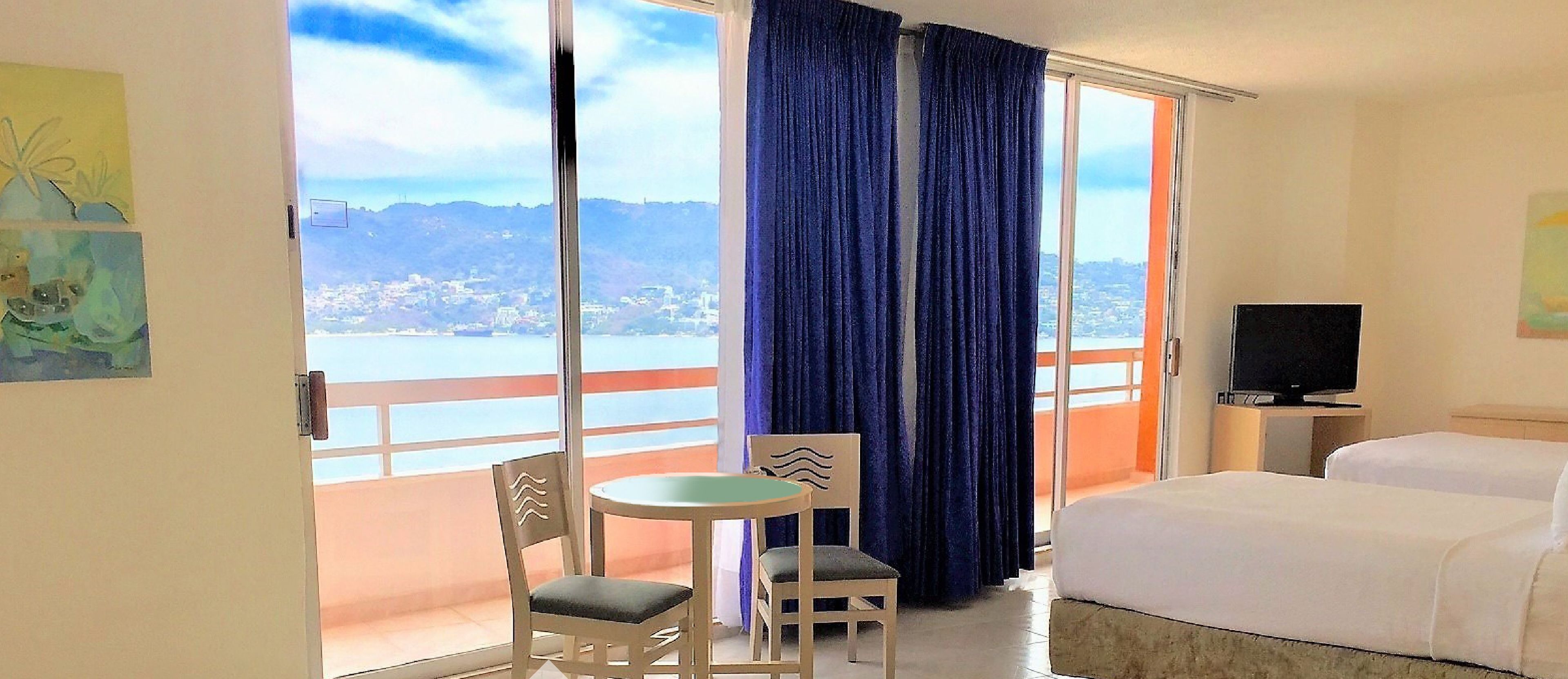 Habitación de hotel con balcón, una cama, televisión y una pequeña mesa con sillas.