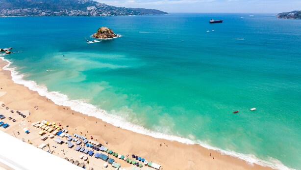 Une plage à l’eau turquoise et limpide, une petite île et un bateau aperçu au loin.