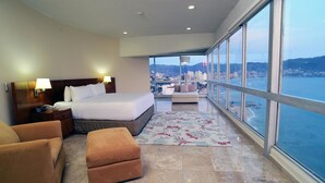 Une chambre d’hôtel avec un grand lit, deux tables de chevet, un canapé, un pouf et une vue sur la mer et le paysage urbain.