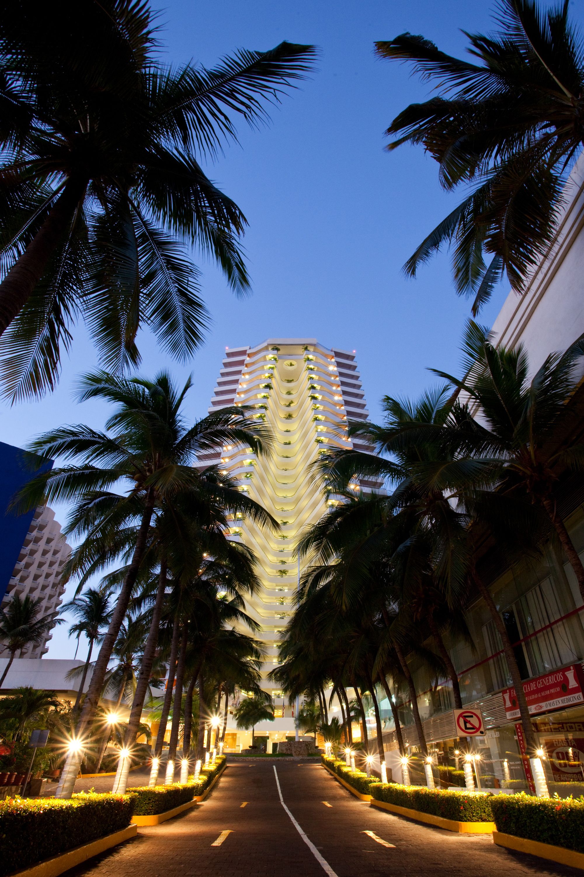 Un hotel con un atrio central, palmeras bordeando la calle y letreros iluminados.