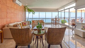 Un vaste espace de vie doté d’une grande fenêtre offrant une vue sur la mer, d’un ensemble de canapés, de fauteuils en osier et d’une petite table agrémentée d’une plante.