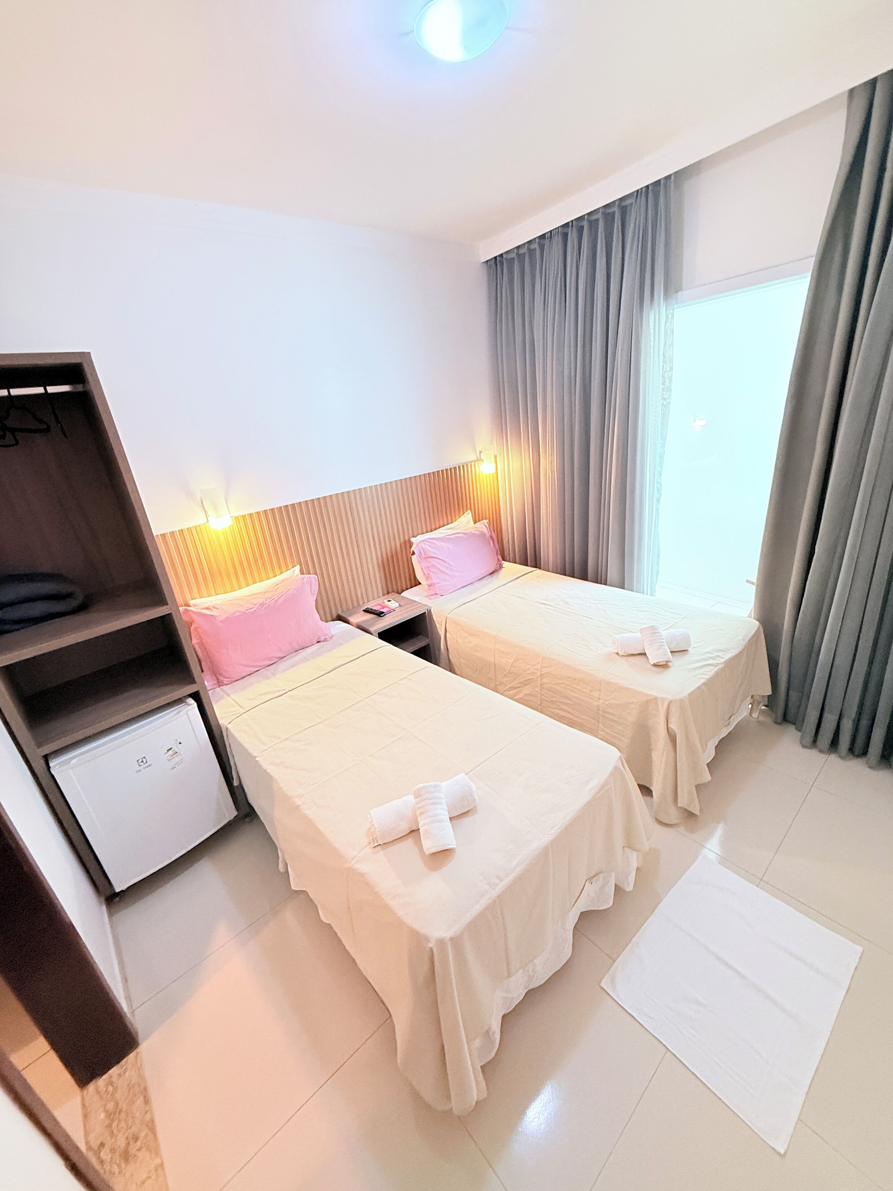A hotel room with two beds, a small fridge, a nightstand, and a window with curtains.