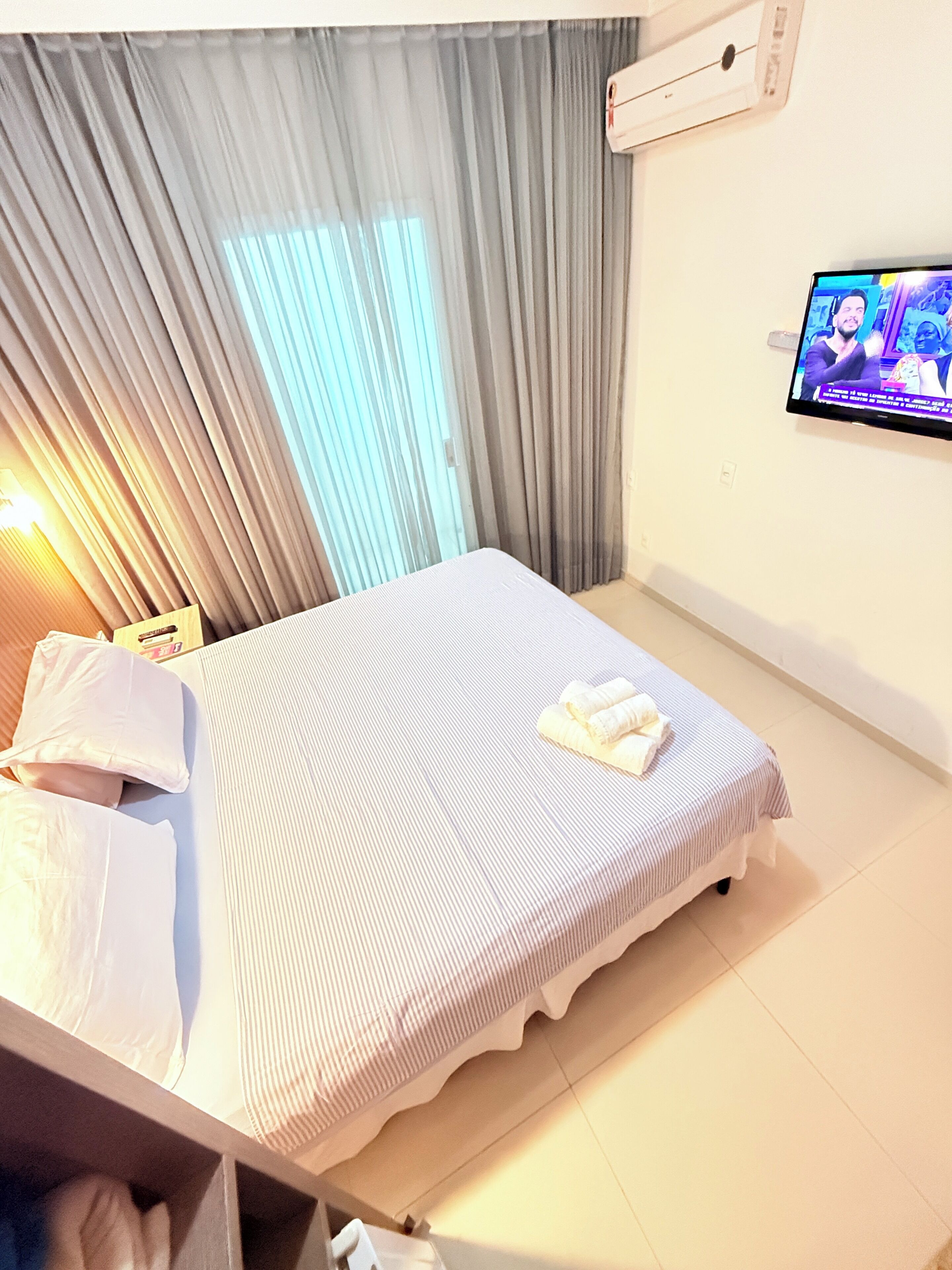 A hotel room with a bed, curtains, an air conditioning unit, and a television on the wall.