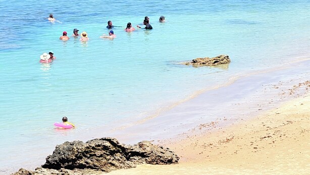 Ein Strand mit klarem, blauem Wasser, badenden Menschen und Palmen im Hintergrund.