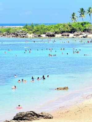 Una playa con agua azul cristalina, gente nadando y palmeras al fondo.