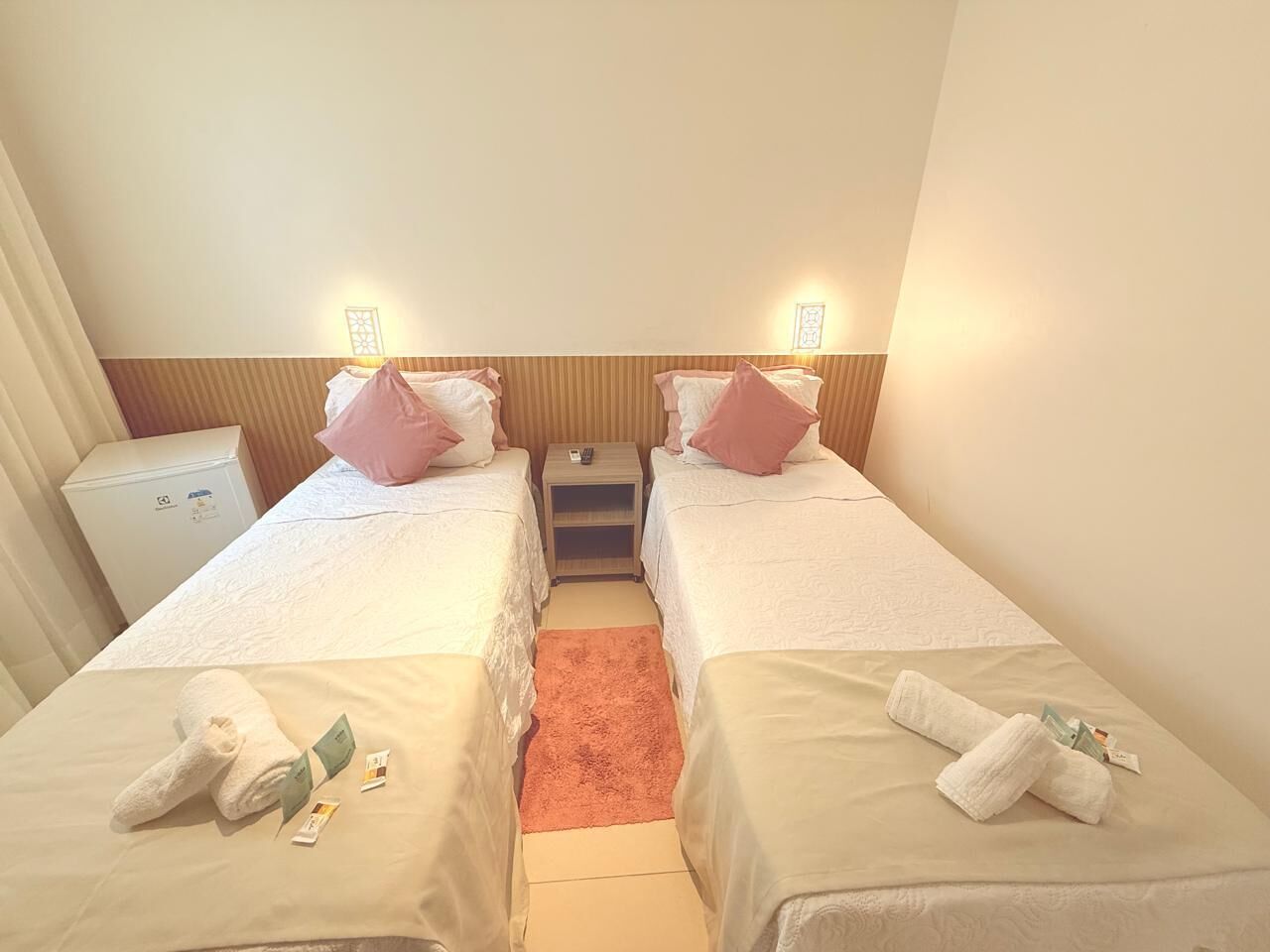 A hotel room with two single beds, each with a pillow and a rolled towel. There is a small table between the beds with a remote control on it.