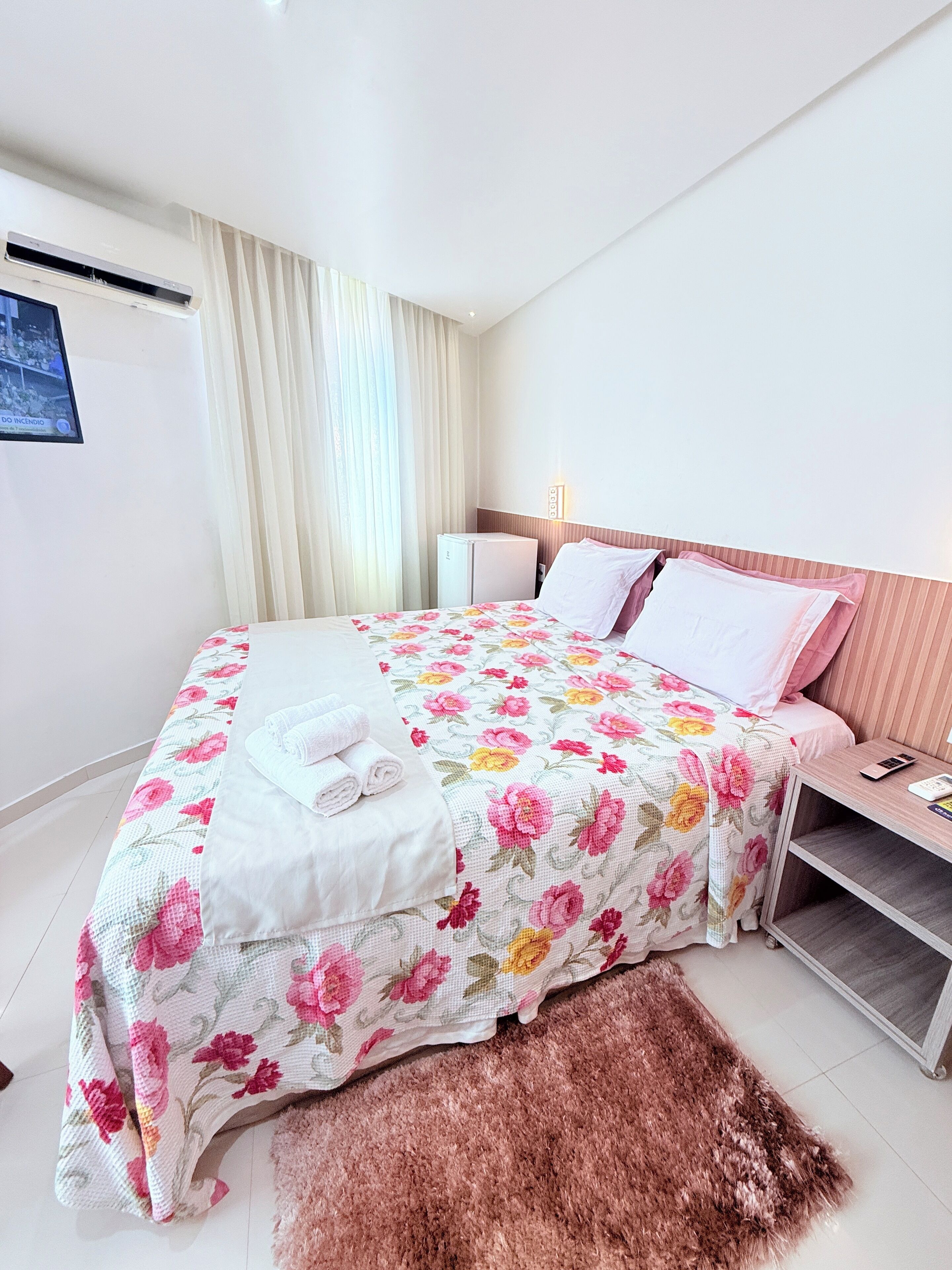 A neatly made bed with floral bedding, a bedside table with a lamp, and a small refrigerator.