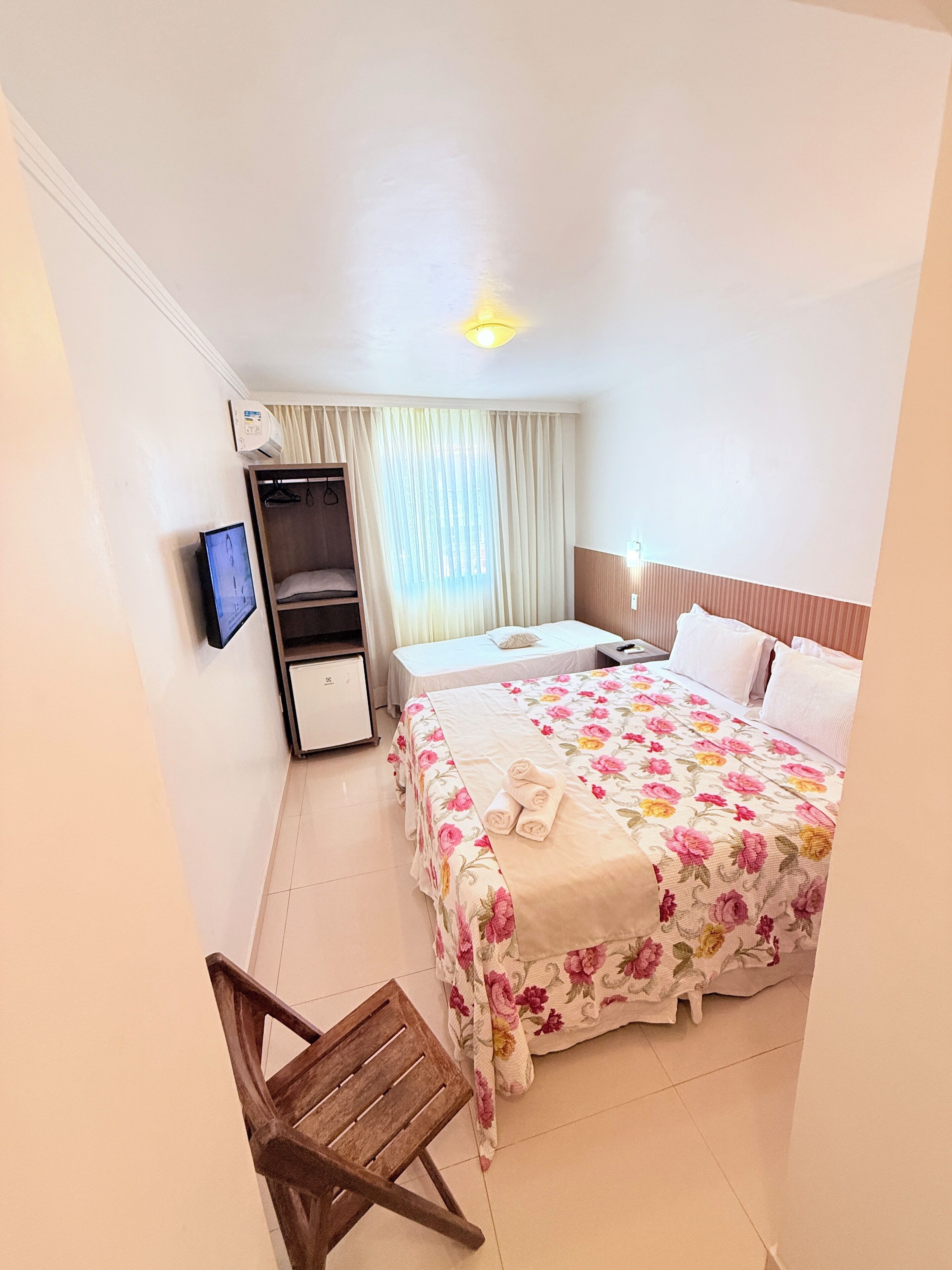 A hotel room with a bed, a wooden chair, a wardrobe, a television, and a window with curtains.