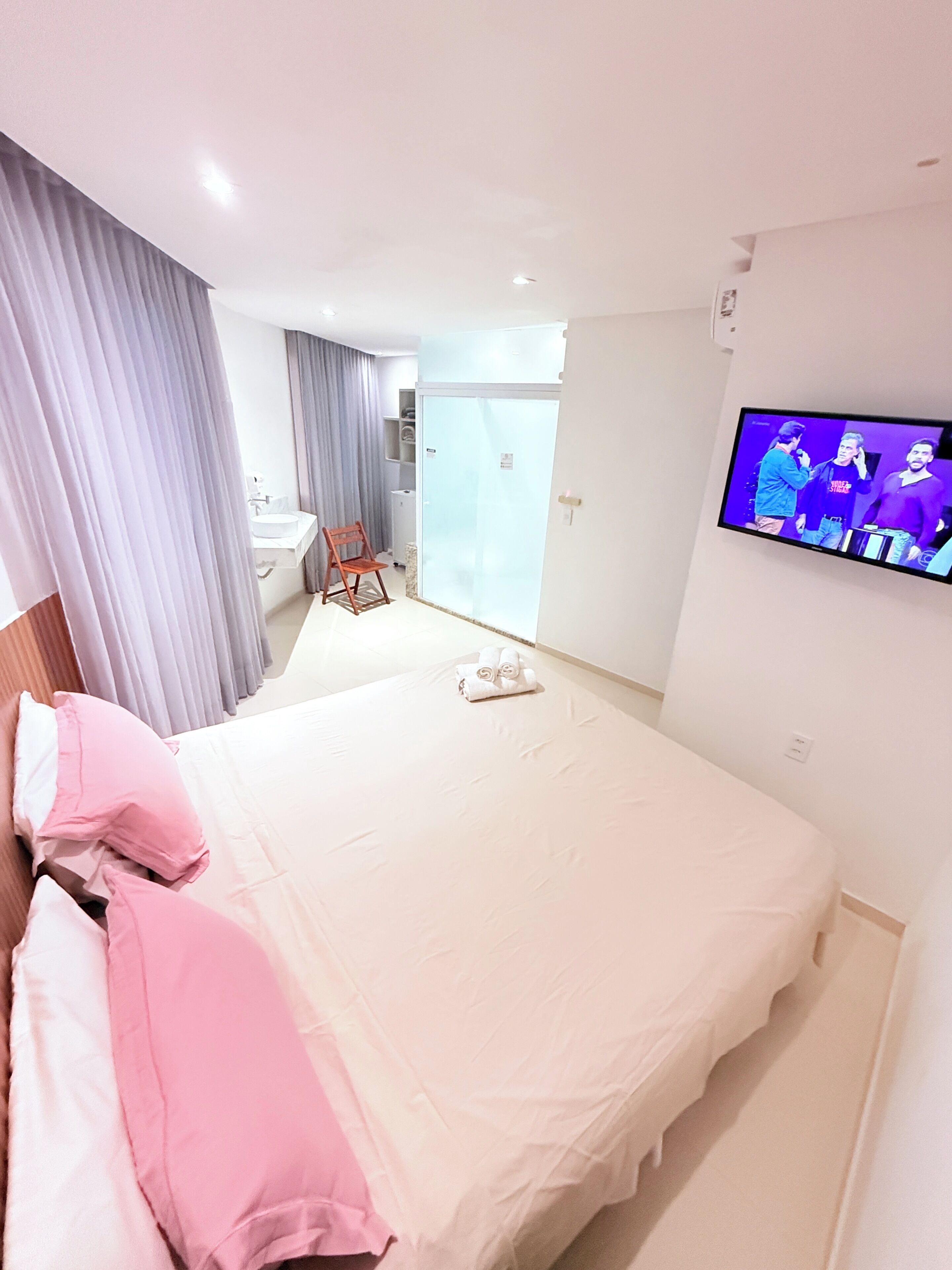 Una habitación de hotel con una cama grande, un televisor montado en la pared, un baño con ducha y lavamanos, y una silla.
