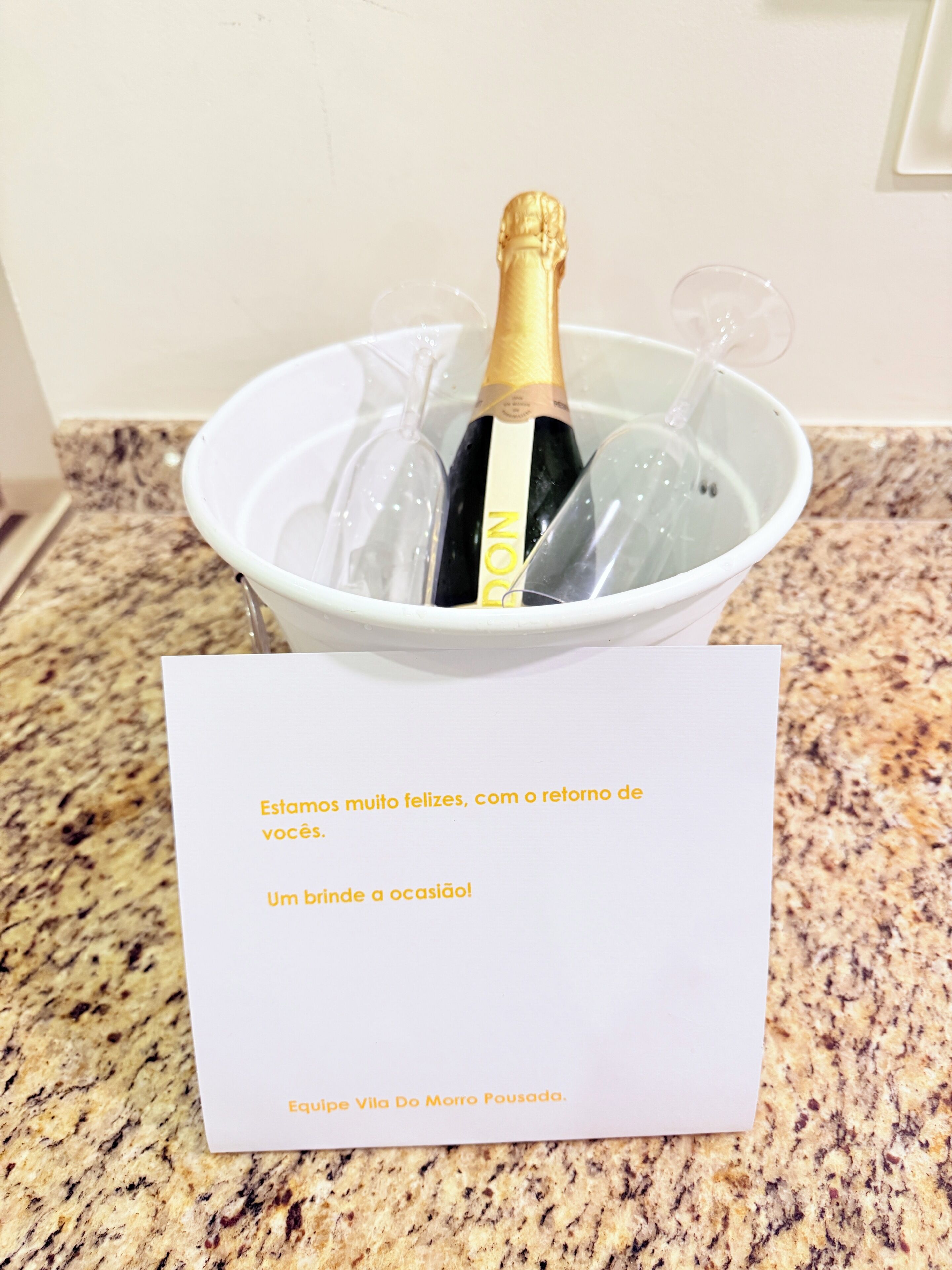 A bucket with a bottle of champagne and glasses, a note with a message, and a granite countertop.