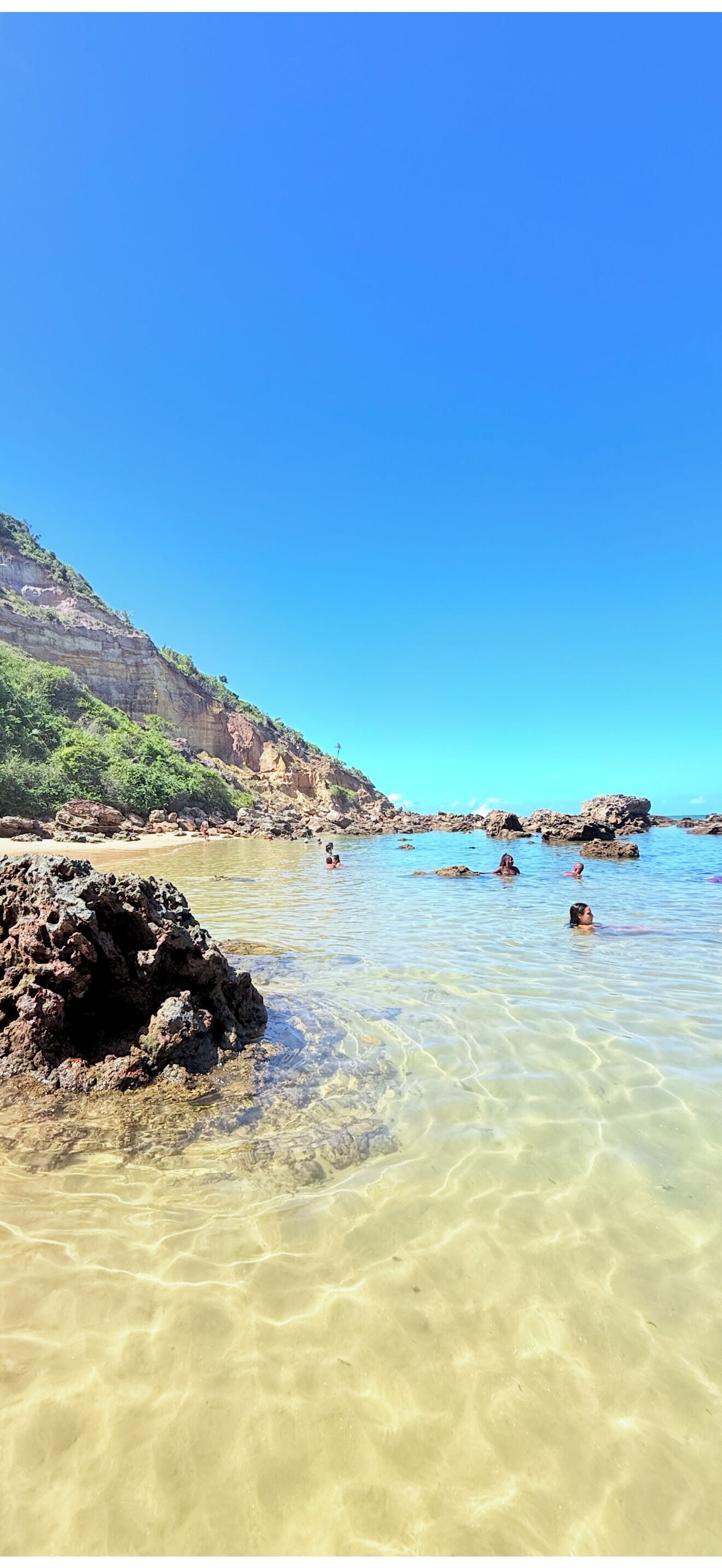 Uma praia com águas cristalinas, formações rochosas e um penhasco ao longe.