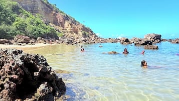 Uma praia com águas cristalinas, formações rochosas e um penhasco ao longe.
