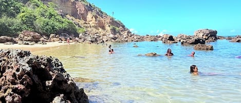 Une plage à l’eau claire, bordée de formations rocheuses et surplombée par une falaise lointaine.