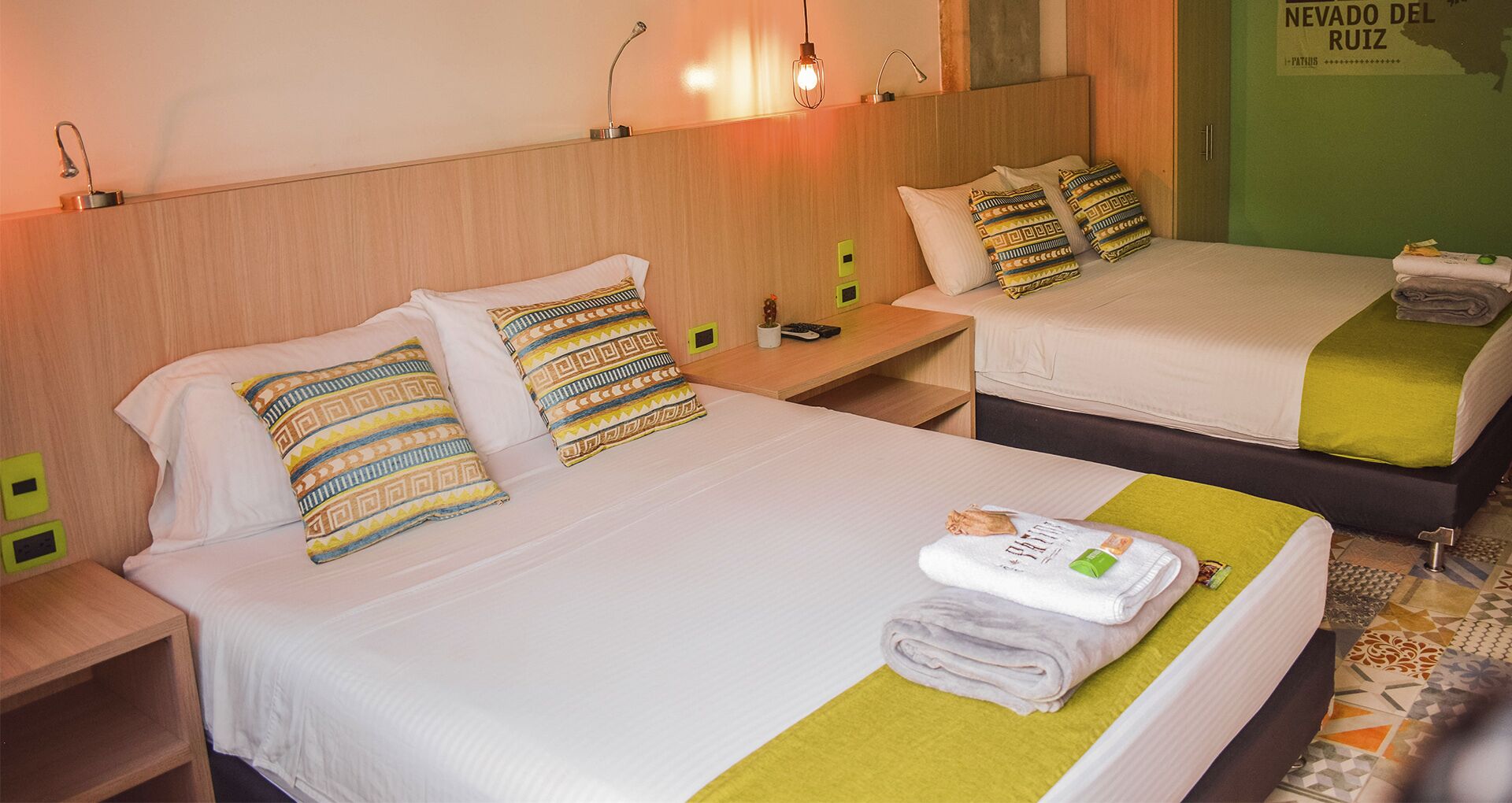 A hotel room with two beds, each with patterned pillows, a small nightstand, and a wall-mounted lamp.