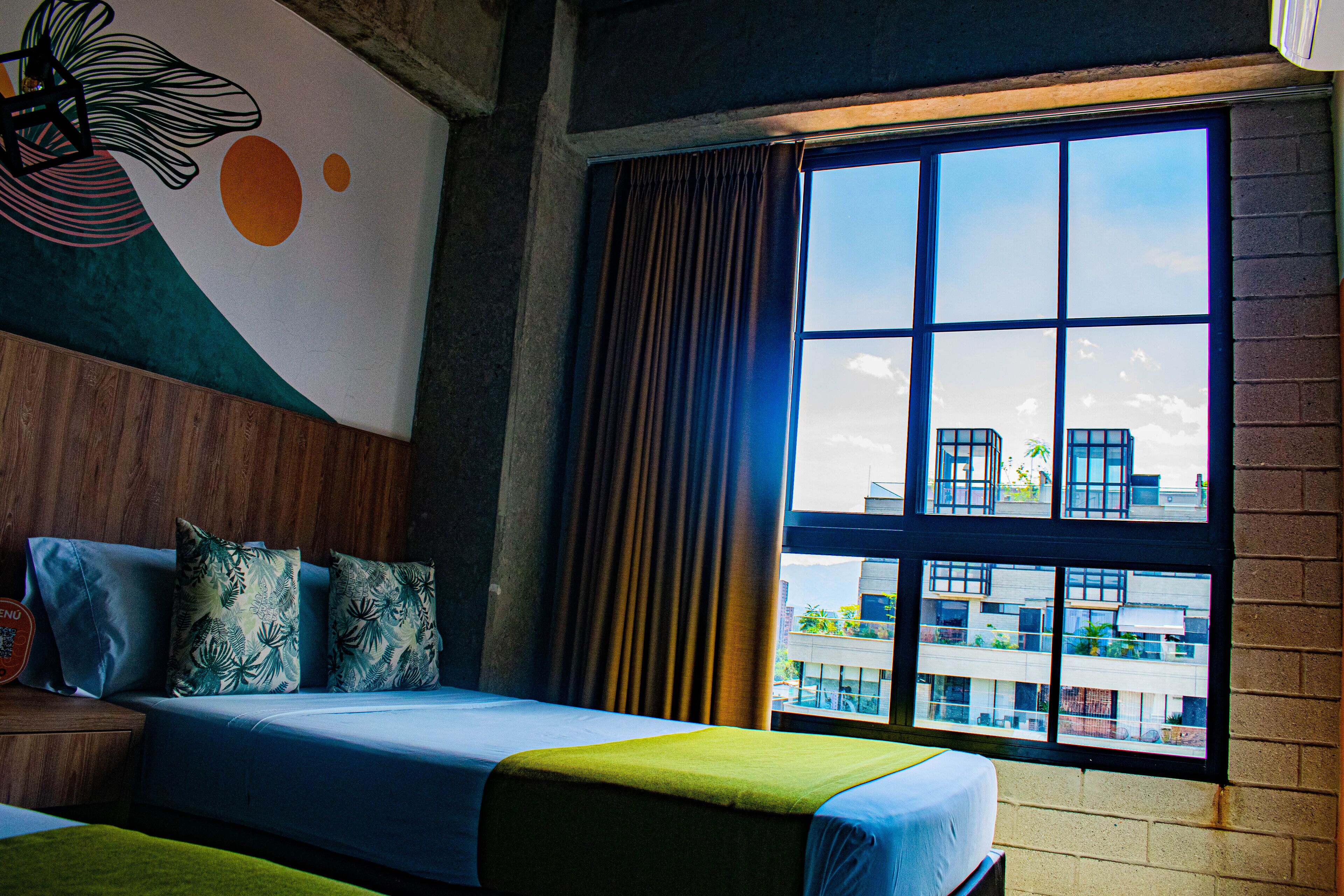 A hotel room with a large window, a bed with a green and blue bedspread, and a wall with a geometric design.